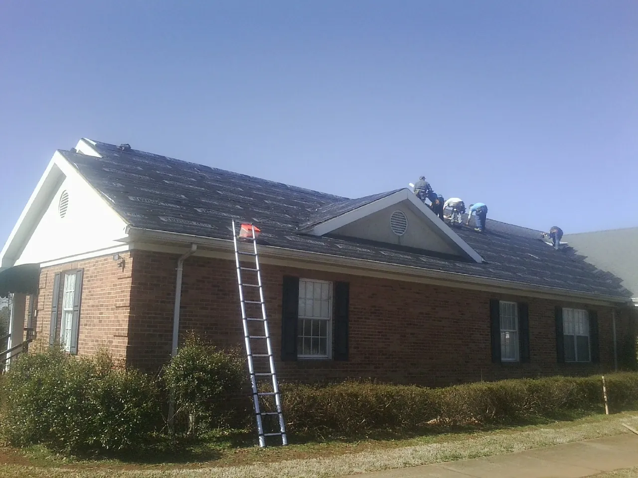 Crew tearing shingles off a brick ranch roof with a ladder set — Metal Roof Repair work by our crew in La Porte City