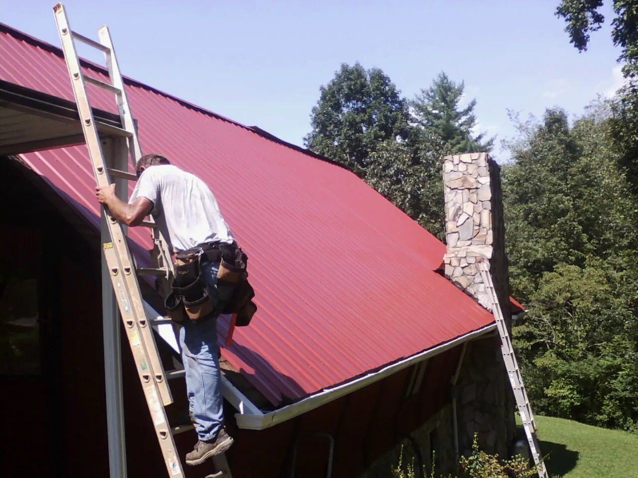 Roofer on a ladder beside a red metal roof and stone chimney — Soffit & Fascia Repair by Roofing Brothers in La Porte City